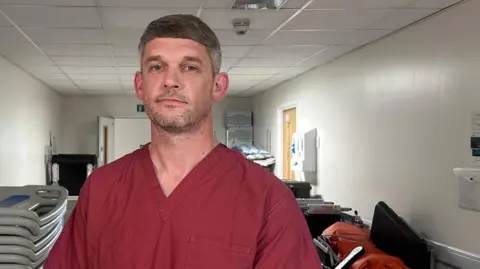 Lee Ellis standing in a hospital corridor. He has short salt and pepper hair and blue eyes. He is wearing burgundy scrubs and is slightly smiling at the camera.