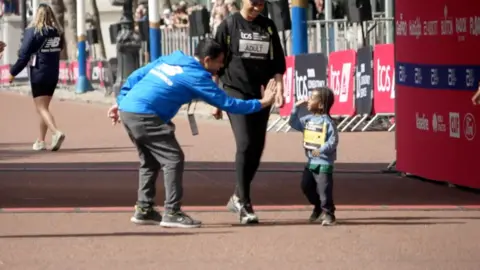Child giving a high-five to a crouching man in a blue New Balance jacket during a race event finish line.