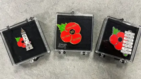 Three custom poppy pin badges lined up in display cases against a grey table. The one on the left has a grey tower clock image with union jacks. The one in the centre is a larger poppy with 'VE DAY' 'VJ DAY' and '1945-2025' written on the petals and '80 in the centre. The third includes sign posts which list Stoke-on-Trent: Longton, Fenton, Hanley, Burslem, Stoke and Tunstall. It also reads '1925-2025' at the top.