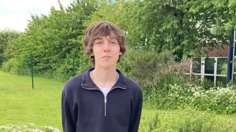 A young man with long brown hair and wearing a blue half-zip fleece stands in front of bushes, trees and grass