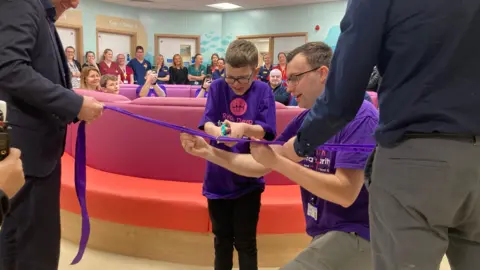 Tim wearing a purple tshirt holds a pair of blue scissors and cuts a long purple ribbon inside the new RD&E children's emergency department. A man is crouched next to him wearing a purple tshirt. Two other men stand holding either end of the ribbon. There is a large group sat on pink sofas in the background.