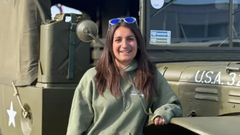A woman in her early 30s with long brunette hair. She is wearing a military green jumper and is stood in front of a World War Two military truck.