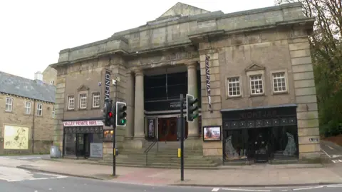 An imposing grey stone building with two tall columns either side of the entrance doors in the middle. Both sides of the building's front have three sets of windows above separate shop fronts. The building sits next to a main road in front of a pedestrian crossing.