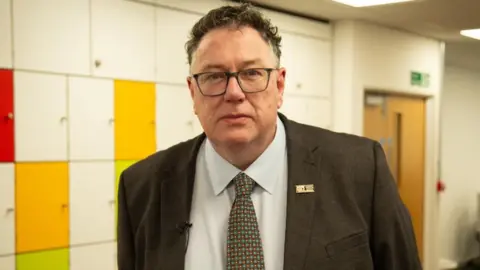 Julian Kennedy is stood in a school hallway. He has short dark hair. He is wearing glasses, a shirt, tie and blazer. He is looking directly at the camera and not smiling.