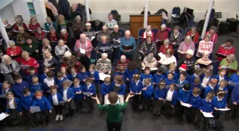 An overhead shot of the choir singing. The children from Combe Down Primary School are at the front of the choir and are wearing blue  uniform, while the Bath Good Afternoon choir are at the back. There is a conductor leading the choir.