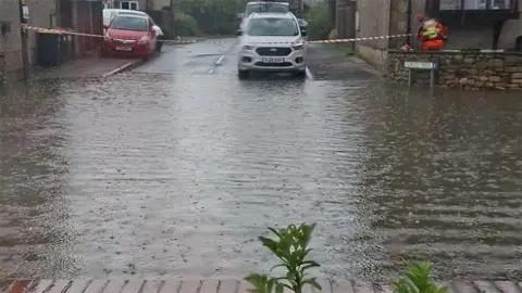 A residential street affected by flooding during wet weather. The photograph is taken from the edge of a driveway or front garden, looking directly across the road. The entire width of the street is covered by standing water, creating a shallow, reflective surface that stretches from one side of the road to the other. Small ripples across the water suggest ongoing rainfall. The water looks to be several centimetres deep.