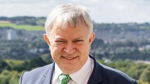 A man with grey hair smiling at the camera. Witha navy suit, white shirt and green tie. In the background there are faded green country fields 