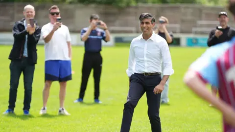 PA Media Rishi Sunak stands in a football pitch with his foot on a football. He wears a white shirt and navy trousers. People stand around him and take photos with their phones. 