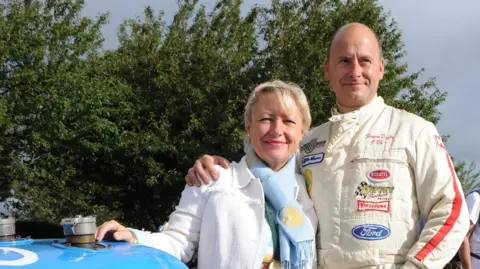 A man with a bald head in a motor racing outfit with his arm round the shoulder of a woman who is wearing a blue scarf and white jacket. They are both smiling. They are standing by an old blue car but you can only make out the bonnet.