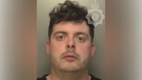 A police custody shot of Luke Williamson, who has short black hair and is wearing a black T-shirt. The wall behind him is grey. 