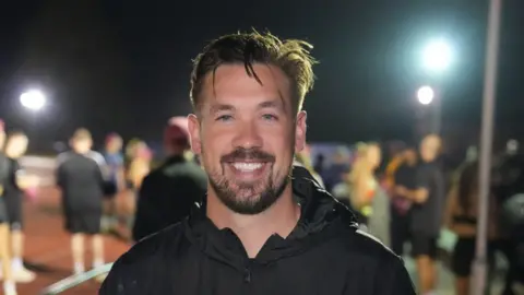 John Fairhall/BBC Luke Chambers is smiling into the camera. He is wearing a black sports jacket. Behind him are other runners on an athletics track. 