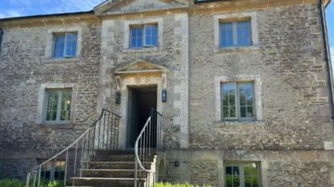The frontage of a Jane Austen-style Georgian brick house with a stone staircase and iron railings leading up to the front door on the first floor. There is a hedge in the foreground, partially obscuring the windows in the basement. The sky is blue above.