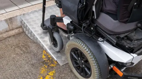 Getty Images A stock image of a powered wheelchair travelling up a short ramp onto a pavement. The wheelchair is black, with grey wheels, and its user is mostly out of shot, with their leg just visible resting on the footplate. The ramp is made out of metal with hatch marks on it to help with traction.