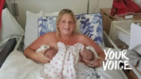 Victoria Oliver lying in a hospital bed, after giving birth to twins. The heads of each baby are in either hand, as she breastfeeds them. A pink spotted white blanket is between the heads. Victoria is smiling at the camera and has blonde hair. The bedsheets are white and there are bars next to the bed. The Your Voice branding is overlaid on the lower right corner of the image