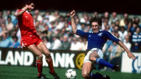 Getty Images Two footballers during a tackle on a pitch. One wears a red shirt, shorts and socks. The other wears a blue shirt, white shorts and blue socks. 