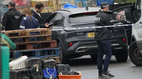 National Crime Agency officers in uniform standing in front of a car and some wooden pallets.