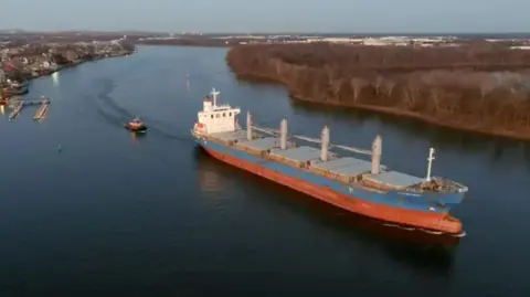 Bill Michaelson The RJN Confidence ship sailing through the water. It is 179 metres long and 28 metres wide. The bottom is painted red and the midsection is blue. It is carrying a lot of cargo on it's deck. 