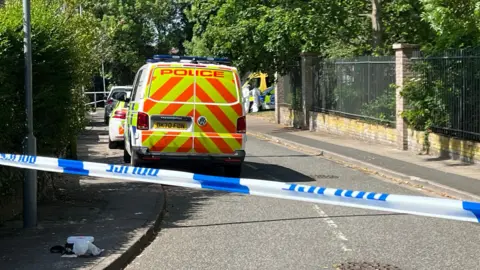 BBC Police vehicles parked on a road behind a police cordon. The road is cordoned off with blue and white police tape. People in white suits can be seen in the distance.
