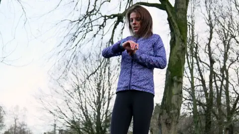 Kristian Johnson/BBC A woman with brown hair in a purple sports jacket stands in a park, looking at her smartwatch.