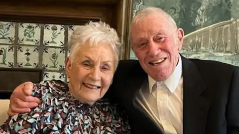 Veronica Ash and her husband are smiling for the camera. Ms Ash is wearing a patterned top and her husband is wearing a black suit jacket and a white shirt.