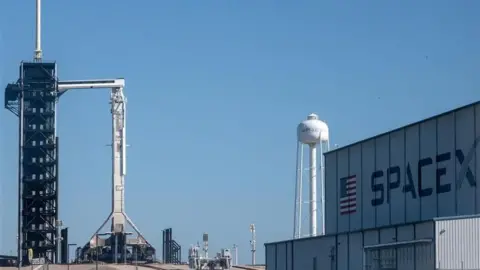 EPA The rocket ahead of launch. Sitting in its stand to prepare to launch, a large building is visible in the near ground with SpaceX written on its side 