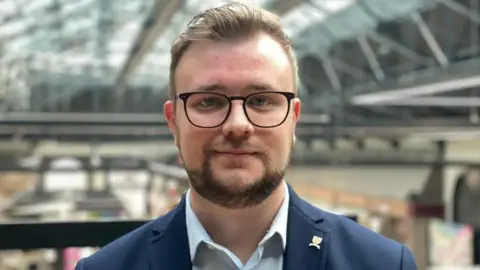 Head and shoulders shot of Joshua Wells inside the market hall. He has a short beard and is wearing a blue suit and glasses. 