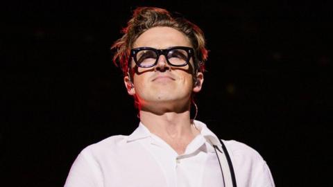 Head and shoulders shot of Tom Fletcher from McFly in a white top and thick black glasses