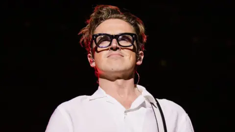 Getty Images Head and shoulders shot of Tom Fletcher from McFly in a white top and thick black glasses