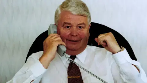 Nationwide Produce PLC handout Bernard O’Malley with grey hair smiling as he holds a telephone to his ear sitting on an office chair wearing a white shirt with a dark red tie and gold tie pin and silver and gold watch