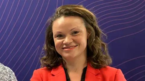 Council leader Bella Sankey has long brown wavy hair and wears a bright red suit jacket over a black top. This photo is taken in a radio studio with a purple background.
