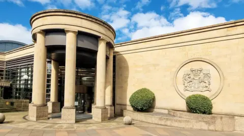 The High Court building in Glasgow is a light brown sandstone building with a large engraved crest on one wall and columns arranged in a circle around the entrance 