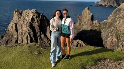 Siobhan Cox Siobhan and Asling are smiling with the ocean and and a cliff edge behind them. Both have dark hair. Siobhan has sunglasses perched on top of her head and is wearing a jersey, jeans and trainers. Aisling is wearing sunglasses, a jersey, shorts and trainers. 