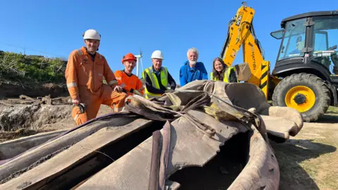 BBC Five people kneel - three wearing hi-vis jackets and hard hats - beside the skull of a whale head. There is a yellow digger on the background.