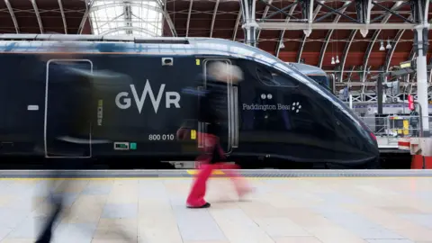 A dark coloured Great Western Railway train sits at London Paddington underneath a rounded roof. Two passengers can be seen blurred as they walk past in opposite directions.