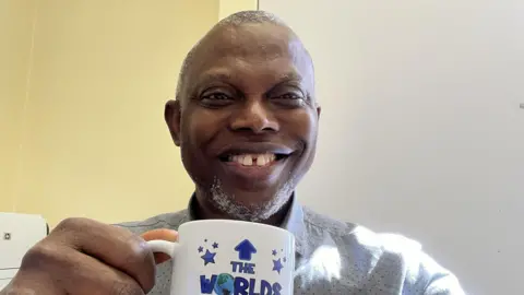 Dumbor Ngaage smiles at the camera as he holds a mug that reads "the world's best professor". He is wearing a white shirt covered in tiny dots. He has a short cropped beard and short grey hair. 