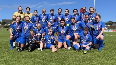 A team of female football players in a blue strip gather in two rows, with some kneeling and sitting while others stand behind. They wear silver medals around their neck which have colourful ribbons.