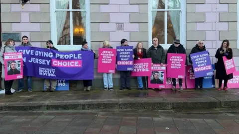 11 people stand in a line against the pink and grey wall of Jersey's States Assembly building. They are holding pink and purple signs and banners. One reads 'Give dying people in Jersey a choice', and another says 'Campaign for dignity in dying'.