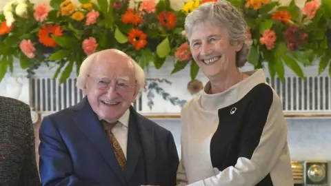 PA Media Michael D Higgins, an older man with balding, white hair, smiles as he shakes hands with Catherine Connolly. He is wearing a navy blue suit, a white shirt and a  patterned tie. Connolly, is also smiling, and is wearing a grey and black suit. She is an older woman with short, grey hair. They are standing in front of a white marble fireplace, with a garland of orange, pink, white and yellow flowers.