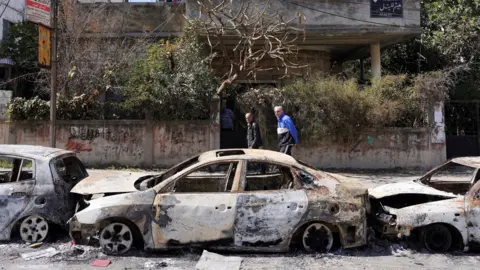 Reuters Two men walk on the pavement past three burned out cars, in front of a building with graffiti on it.