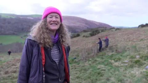 BBC Woman with long, light brown, curly hair is standing on a hillside. She's wearing a bright pink beanie hat and a dark coloured coat. She's smiling at the camera. In the background you see three people on the hillside. Two are working on creating a dam from willow stakes. 