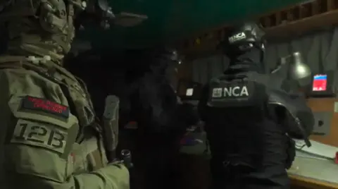 Ministry of Defence Screengrab from video footage depicting three people from behind. The person closest to the camera is in green tactical gear with a patch reading "Royal Marine Commandos", while the other two are in black gear. The back of the uniform reads "NCA National Crime Agency". They are in a dimly lit room.