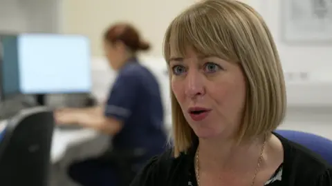 Diane Stafford, has bobbed blonde hair and is wearing a blue uniform