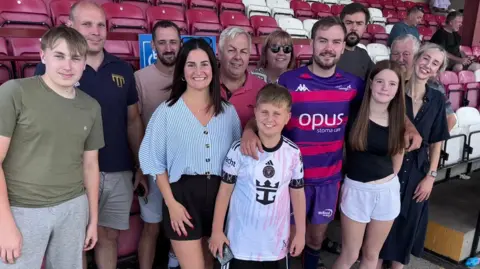 Group of 12 people smiling at the camera in the stands of the Batley rugby league stadium