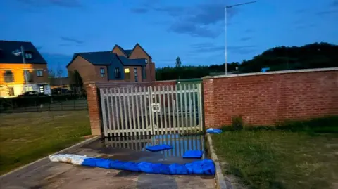 Coral Aimee Blue and white sandbags in front of metal gates. There are houses in the background.