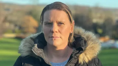 Micala Trussler looks at the camera. She has blond hair tied pack in a ponytail and is wearing a thick black coat with a fur lined hood. Behind her is a green field and trees.