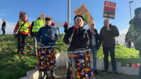 BBC A recent protest. It shows a man and a woman with large drums in the foreground and other protestors holding signs in the background. The signs say "save Adlington" and "no to Adlington new town"