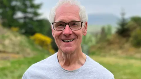 A grey-haired man with glasses, and wearing a grey T-shirt, is smiling at the camera with a woodland behind him