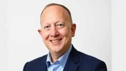 Hertsmere Borough Council A studio portrait of Jeremy Newmark with very short grey hair wearing a dark blue suit jacket and light blue shirt, seated against a plain light-grey background and facing the camera.