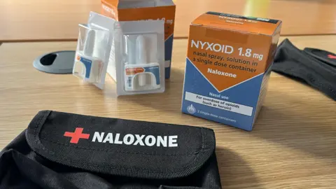 Blue and orange naloxone boxes and white nasal sprays in packaging on a wooden table, with a black velcro kitbag in the foreground featuring the word 'naloxone' on it