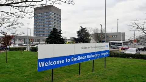 A sign saying "Welcome to Basildon University Hospital" on a grass verge. A hospital tower block and lower level buildings are behind it.